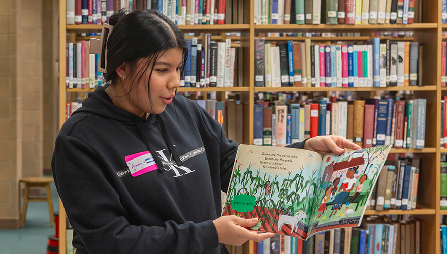 A female student standing in front of library shelves holding a children's book up to show pictures