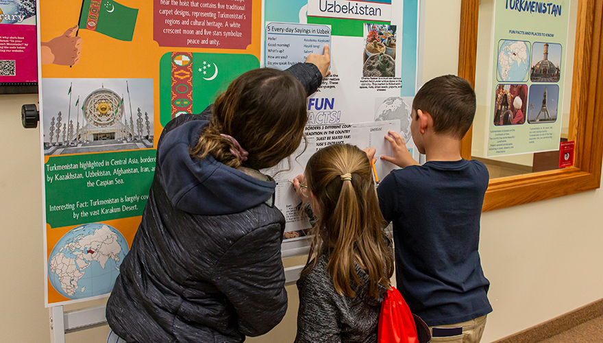 A student teacher pointing at posters on a bulletin board with one boy and one girl holding papers against the board and writing
