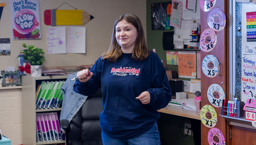 A female college student standing at the front of an elementary school classroom talking to students