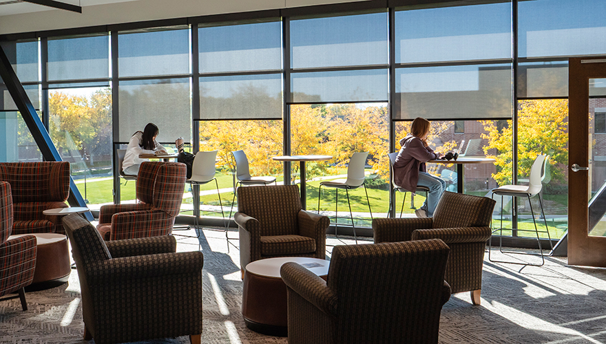 Two students sitting at high top tables studying in front of large windows showing a bright fall day