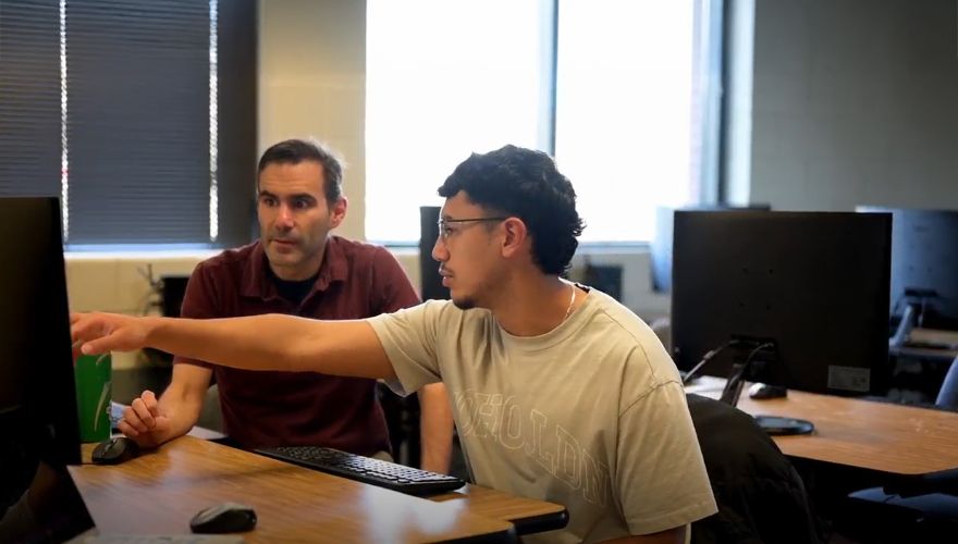 Male student wearing glasses pointing at a computer screen sitting next to a male professor who is looking at the screen in a classroom filled with monitors on tables