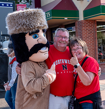 The Trapper mascot wearing traditional trapper clothing posing with a man and a woman on the street