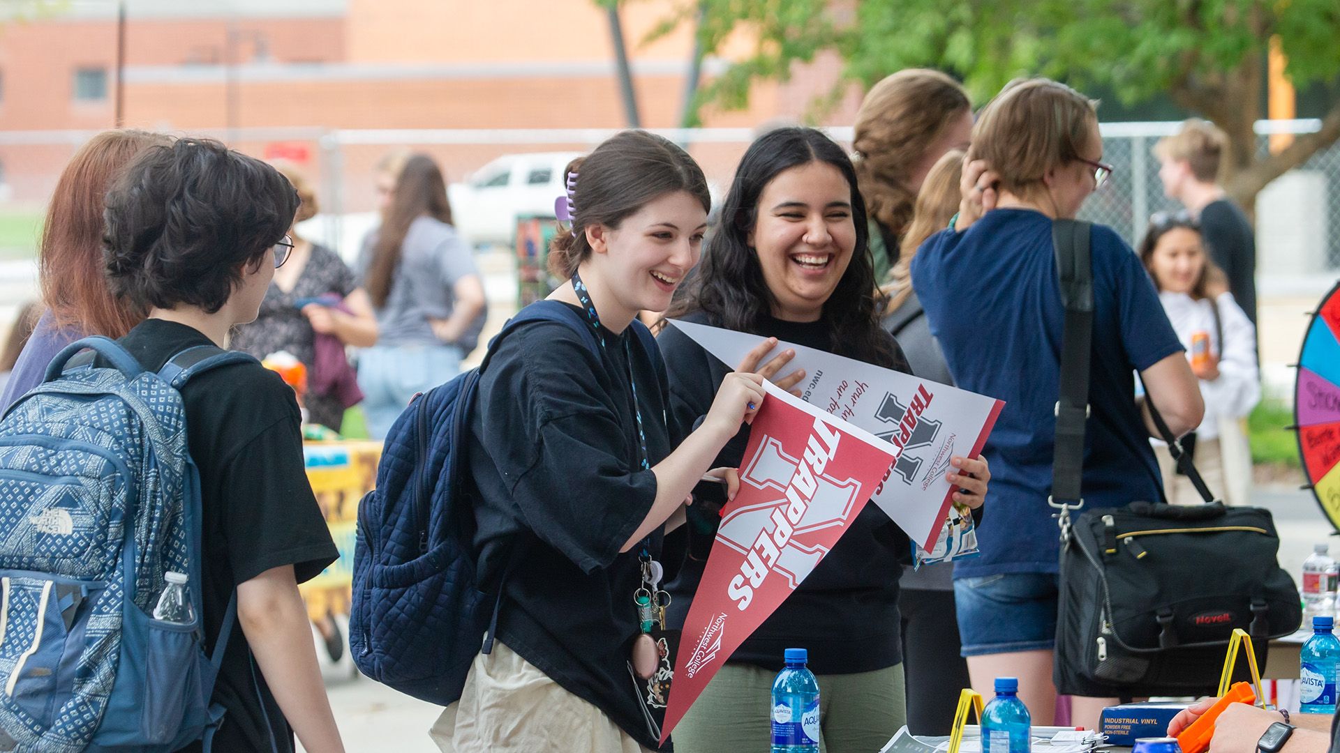 Two smiling female college students standing at a table outside holding Northwest College pennants