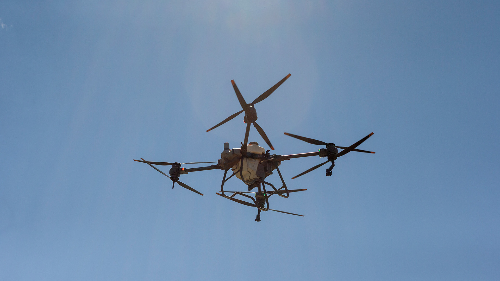 A large drone hovering against a blue sky with a slight lens flare behind it