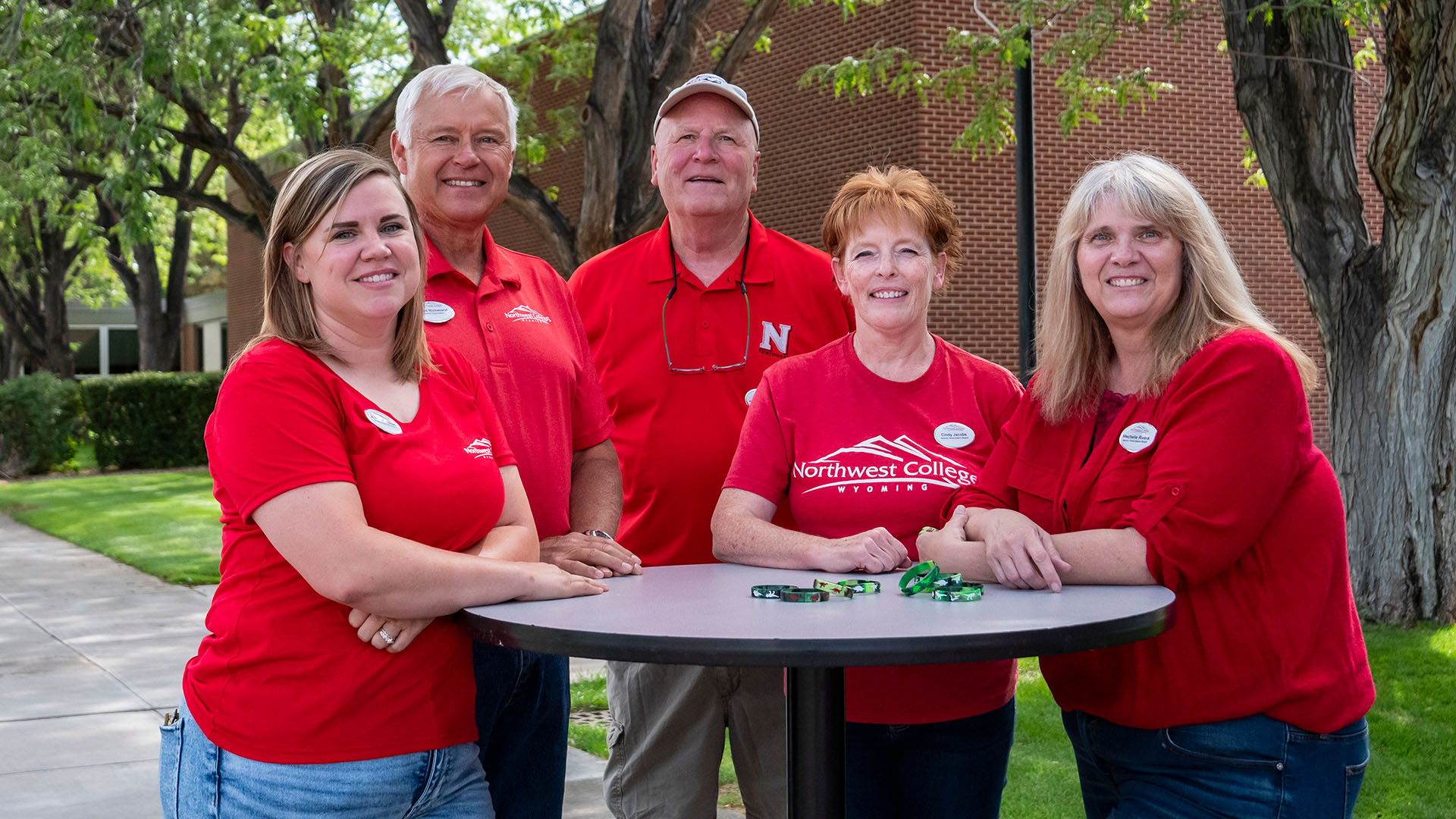 Three women and two men in various red shirts standing around a high top circular table outside under trees