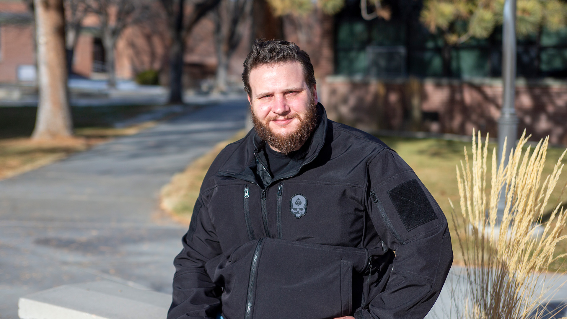 Photo of a bearded man wearing a black jacket, sitting on a bench in sunlight with trees and red brick buildings in the background