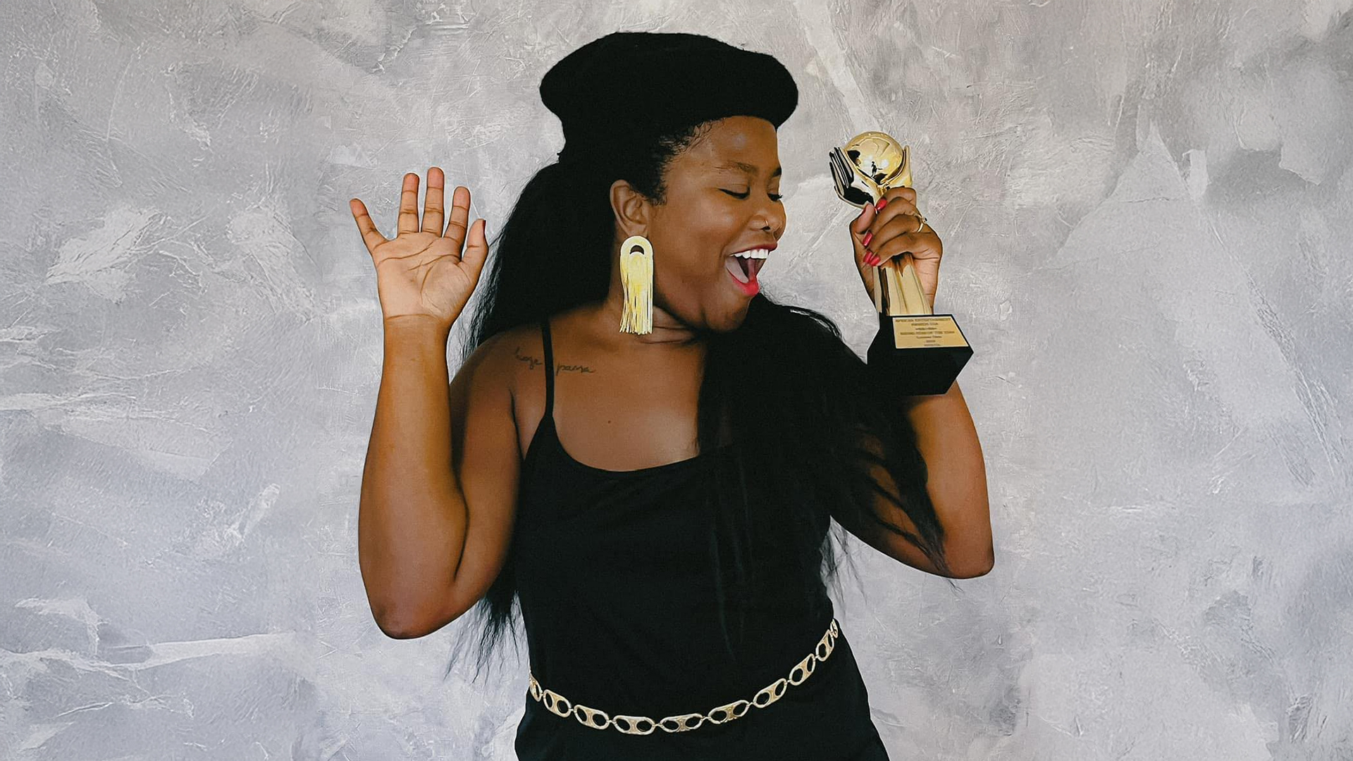 Singer Luciane Dom holding a trophy, smiling and dancing against a gray background