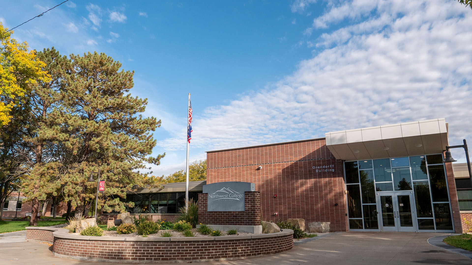 The front of the Orendorff Building with red brick front, signage, and flag pole, floor to ceiling windows around the doors, all against a partly cloudy blue sky and trees on the left