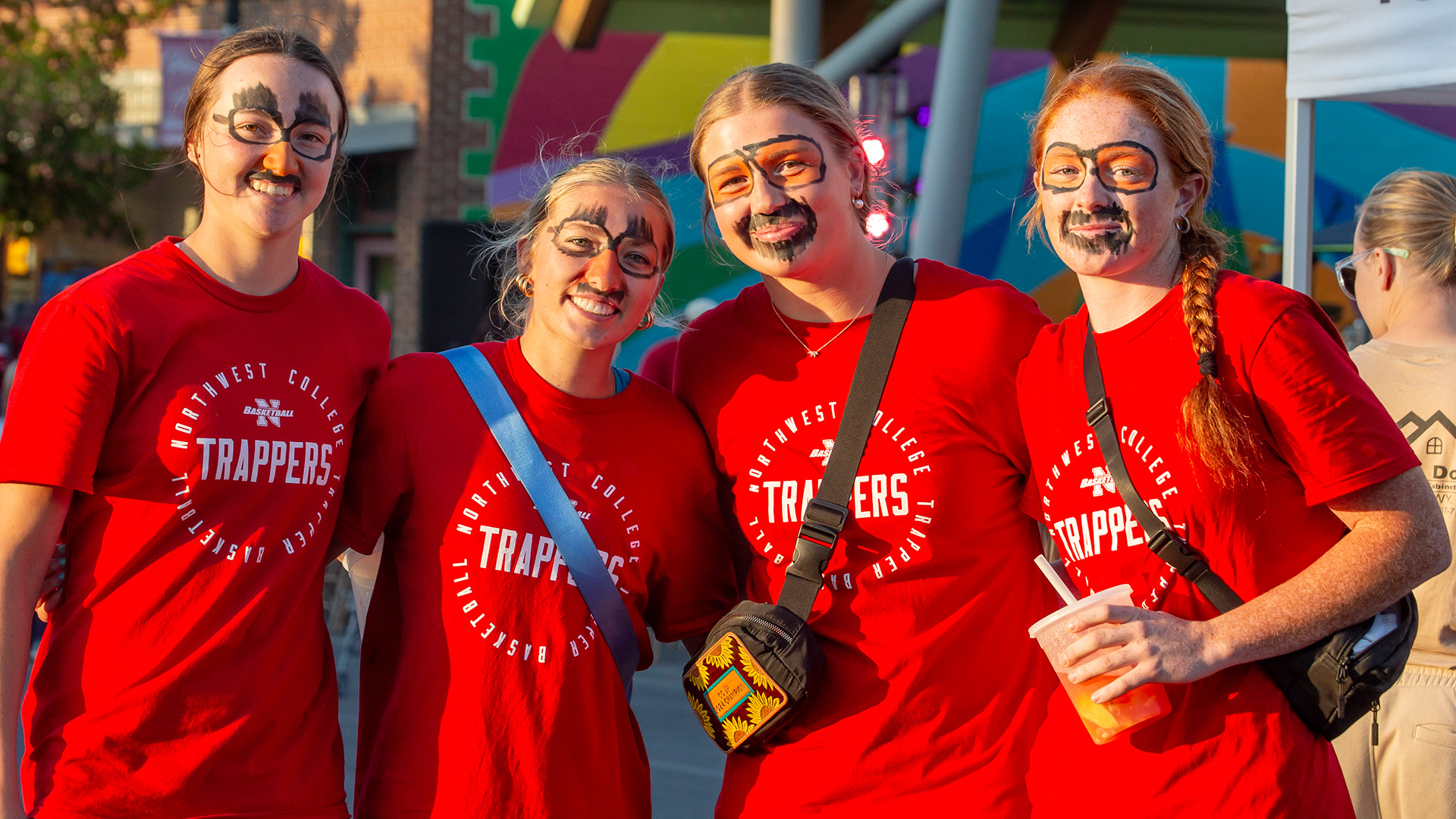 Four female college students with painted faces wearing red Northwest College Trapper shirts in late afternoon sunshine