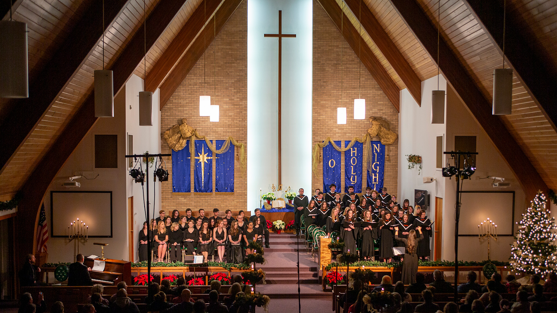 Dimly lit church sanctuary decorated for Christmas with a choir singing in front of purple banners and a large cross