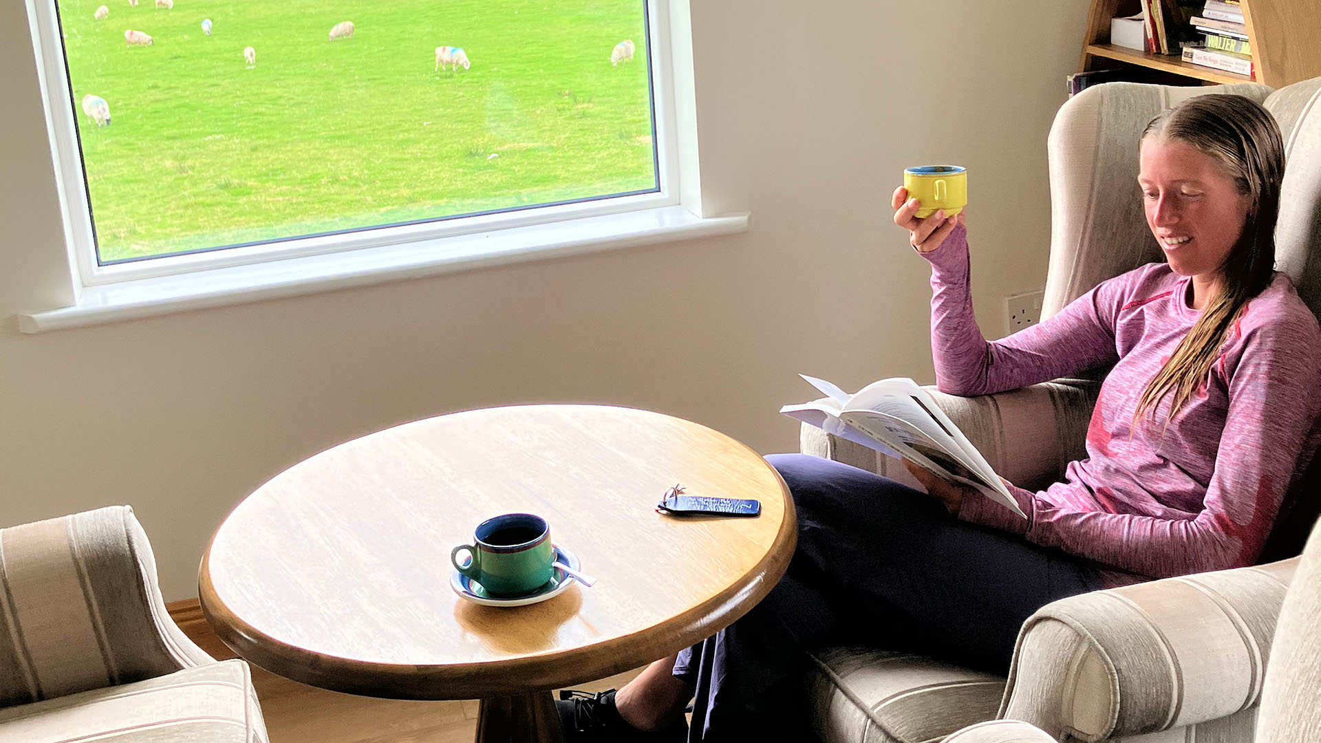 A young woman sitting in a comfortable chair beside a small circular table, reading a book and holding a cup of coffee with a window in the background showing a green field filled with sheep, and a bookshelf behind her